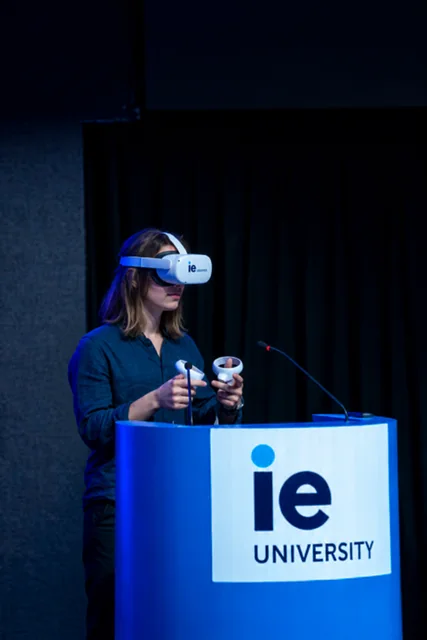 A participant using a VR headset and controllers behind an IE University lectern.